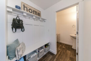 Mudroom interior with a view of a sink on the other room. There is a shelf with decorative board on top and a bag hanging on one of the hooks and a built in bench with linen storage baskets below.